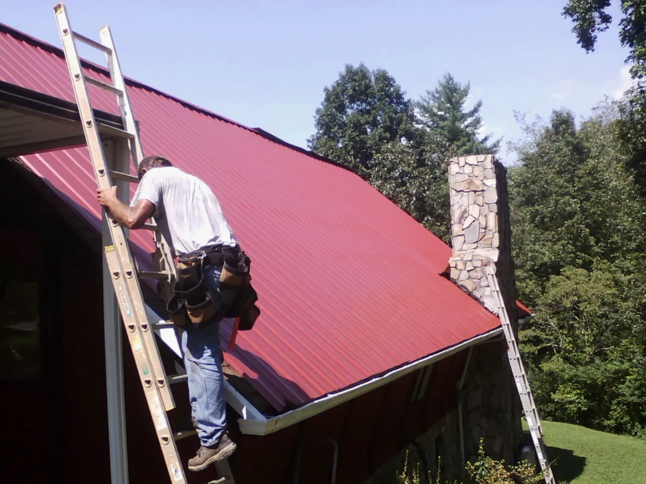 Roofer on a ladder beside a red metal roof and stone chimney — Soffit & Fascia Repair by Roofing Brothers in Matherville